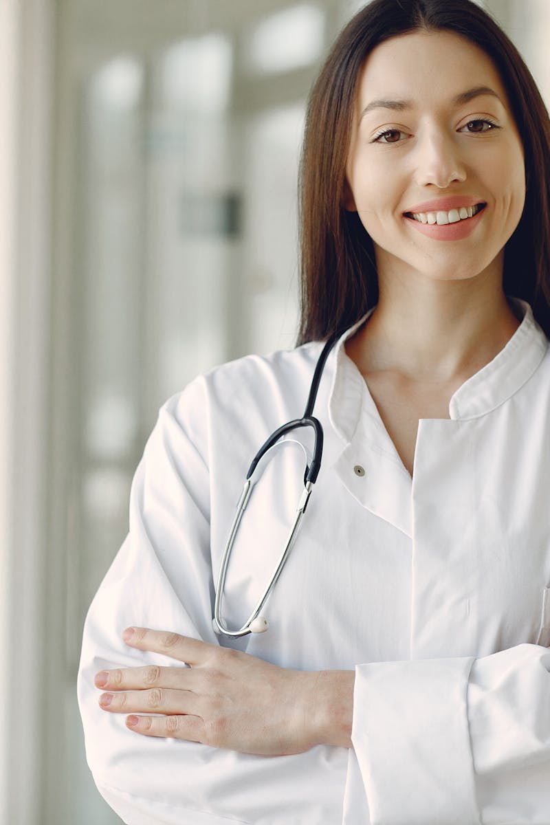 Crop cheerful ethnic female doctor in medical uniform and stethoscope holding hands crossed standing in modern clinic corridor with glass wall
